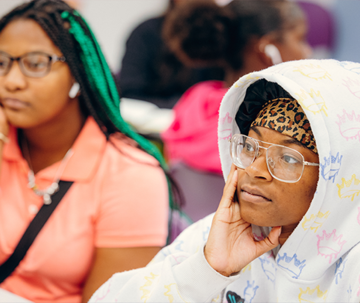 Two students attentively listen in a classroom. The student in the foreground wears a patterned hoodie and glasses, resting their chin on their hand. The atmosphere is focused and thoughtful.